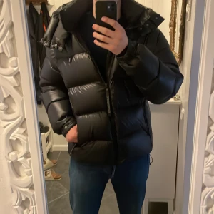 Svart pufferjacka från J.Lindeberg - Säljer fetaste svarta pufferjacka från J.Lindeberg i utomordentligt skick. Jackan är perfekt för vintern med sitt tjocka material och långa ärmar. Den har en dragkedja framtill och en hög krage för extra värme. Passar perfekt för kalla dagar