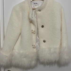 Vit pälsjacka från SJM - Superfin vit pälsjacka från SJM med fluffiga detaljer vid ärmslut och nederkant. Jackan har dekorativa knappar med pärldetaljer och en quiltad insida för extra värme. Perfekt för att glamma upp vinteroutfiten! ❄️✨