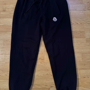 Svarta mjukisbyxor från Moncler - Säljer ett par svarta mjukisbyxor från Moncler i mycket bra skick. De har en bekväm passform med resår och snörning i midjan. Perfekta för en avslappnad dag hemma eller en snabb tur till affären. Moncler-loggan syns tydligt på sidan. Passar perfekt för höst och vinter! Byxorna är i storlek M men passar både S/M.