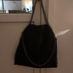 Svart axelväska med kedjedetaljer - Snygg svart axelväska i syntetmaterial med coola kedjedetaljer. Perfekt för en utekväll eller som en stilren vardagsväska. Väskan har en enkel och elegant design som passar till det mesta. Kedjan ger en extra touch av stil och gör den lätt att bära.