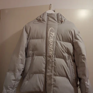 Grå pufferjacka från Trapstar - Säljer en snygg grå pufferjacka från Trapstar i storlek S. Jackan har en stor logga på både fram- och baksidan, vilket ger den en cool look. Den är perfekt för kalla dagar med sin tjocka vaddering och har en dragkedja framtill. Huvan är justerbar för extra skydd mot väder och vind. Typ helt ny.