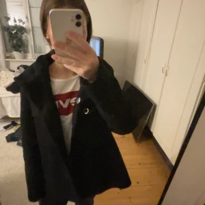 Svart kappa med knappar - Säljer en stilren svart kappa i mycket bra skick. Den har långa ärmar och knappar framtill. Perfekt för höst och vinter, och funkar både till vardags och fest.  den är superbekväm att ha på sig. Passa på att köpa denna snygga kappa!