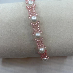 Rosa armband med pärlor - Ett superfint armband med rosa flätad design och vita pärlor. Perfekt för att ge en söt och elegant touch till din outfit. Armbandet har en guldfärgad låsning som gör det enkelt att ta på och av. Passar perfekt till både vardag och fest!