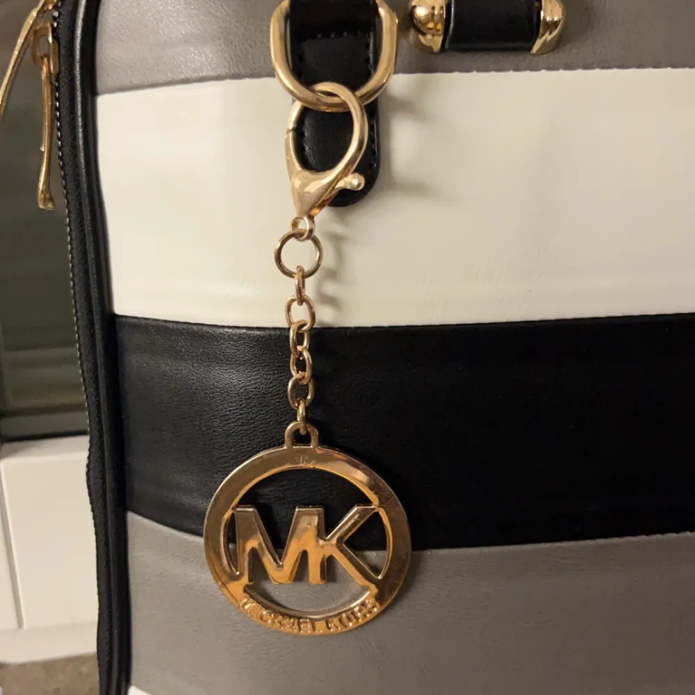 Snygg handväska  från Michael Kors i svart, vit och grå. Väskan har ett elegant randigt mönster och är tillverkad i skinn. Den har en praktisk dragkedja och ett dekorativt guldfärgat MK-hänge. Perfekt för både jobb och vardag! Måste sälja för att ska flytta . Väskor.