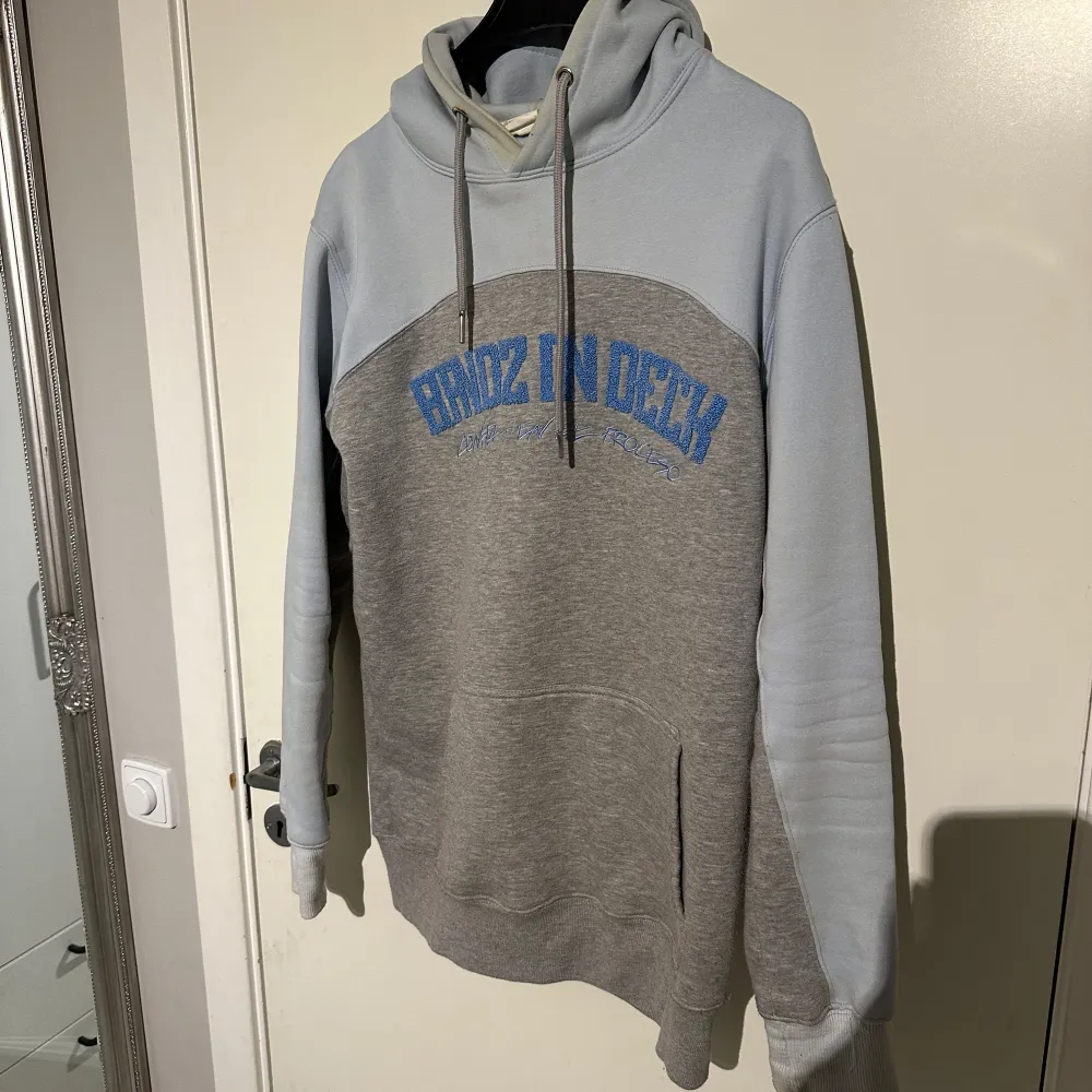 Säljer en snygg grå och vit hoodie med blå text på bröstet. Den har en stor ficka fram och en bekväm huva. Perfekt för en avslappnad stil och passar bra till jeans eller joggers. Skön att ha på sig under kyliga dagar.. Tröjor & Koftor.