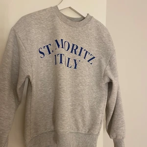 Grå sweatshirt från Gina Tricot - Säljer en snygg grå sweatshirt från Gina Tricot i storlek XS. Den har texten 'ST. MORITZ ITALY' i blått på framsidan. Perfekt för en avslappnad stil och passar bra till både jeans och leggings. Tröjan är i mycket bra skick och supermjuk!