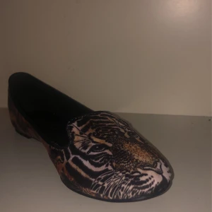 Leopardmönstrade ballerinaskor med tigermotiv - Säljer ett par unika ballerinaskor från TIAMO i storlek 37. Skorna har ett coolt leopardmönster med ett iögonfallande tigermotiv på tån. Perfekta för att ge din outfit en vild touch! De är i bra skick och redo för nya äventyr.
