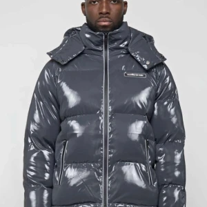 Grå glansig pufferjacka - Säljer en snygg grå pufferjacka med glansig finish. Jackan har en hög krage och en praktisk huva för extra skydd mot kyla. Den är perfekt för vintern och har en dragkedja framtill. Jackan är i mycket bra skick och passar perfekt för kalla dagar.