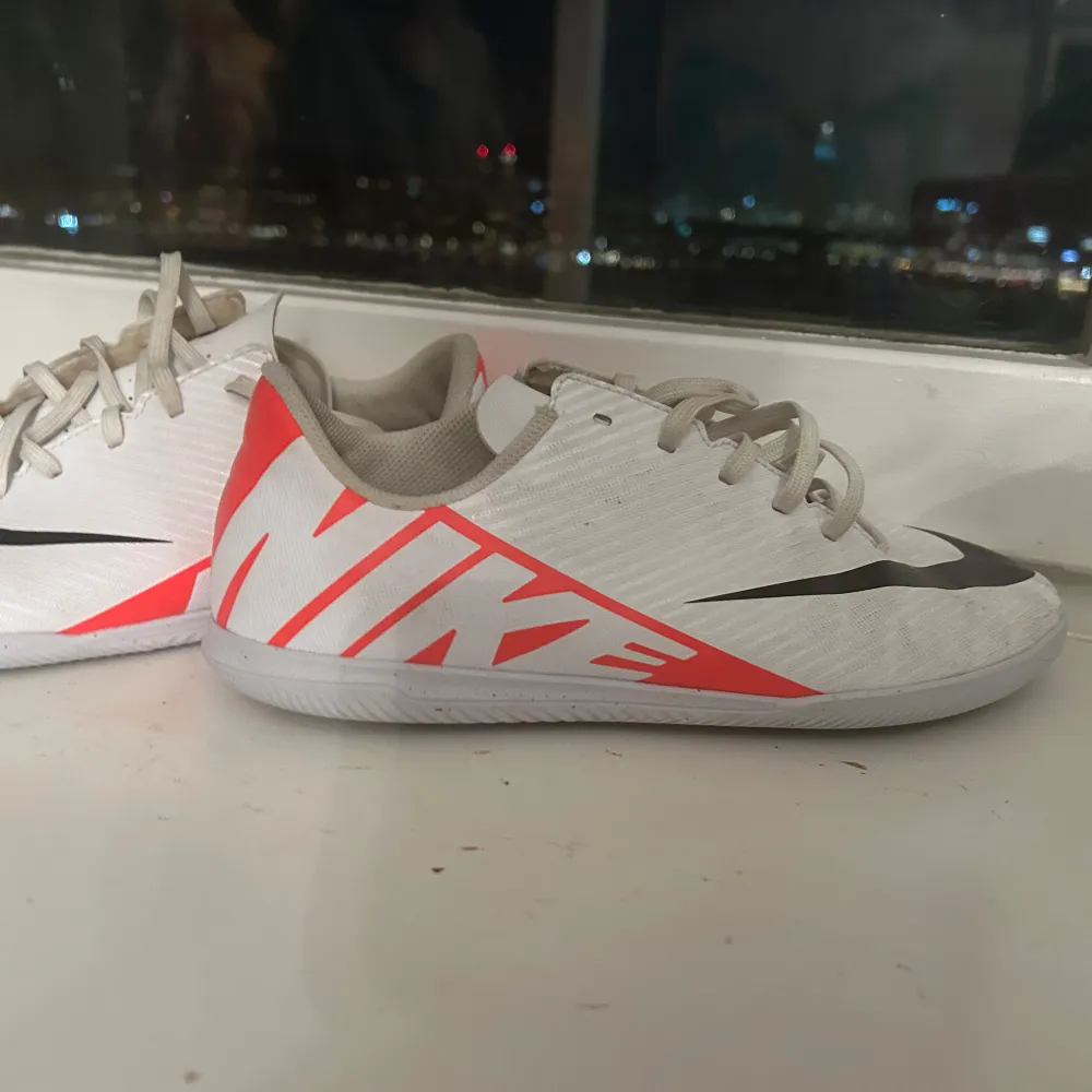 Säljer ett par vita Nike fotbollsskor i mycket bra skick. De har en snygg design med svarta och röda detaljer samt snörning. Perfekta för inomhusfotboll eller träning. Skorna är lätta och bekväma, vilket ger bra kontroll och fart på planen. Använd bara en Sasöng. Skor.