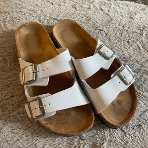 Vita sandaler med spännen - Säljer ett par vita sandaler i mycket bra skick. De har två justerbara remmar med silverfärgade spännen och en bekväm fotbädd. Perfekta för sommardagar och avslappnade stunder. Passar till både strand och stadspromenader.