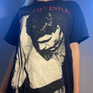 Svart Joy Division t-shirt - Säljer en svart t-shirt med Joy Division-tryck i rött och vitt. Den har en cool vintage-look med ett stort grafiskt tryck på framsidan. Perfekt för dig som älskar bandtröjor och vill ha en avslappnad stil. Passar bra till jeans eller shorts för en casual look.