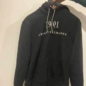 Svart hoodie från Armani Exchange - Säljer en snygg svart hoodie från Armani Exchange med vit text på bröstet. Den har en klassisk känguruficka och justerbar huva med snören. Perfekt för en casual look. Passar till alla tillfällen!