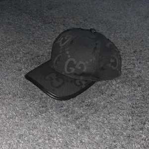 Svart keps med GG-mönster - Snygg svart keps med GG-mönster, perfekt för att ge din outfit en lyxig touch. Kepsen har en klassisk design med böjd skärm och justerbar passform. Materialet känns slitstarkt och bekvämt, vilket gör den idealisk för både vardag och fest.