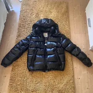 Säljer en snygg svart pufferjacka från Moncler i mycket bra skick. Jackan har en glansig finish och en praktisk huva. Den är perfekt för kalla dagar och har en dragkedja framtill. Moncler-loggan syns tydligt på armen. Passar perfekt för vintern!