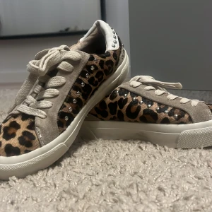 Leopardmönstrade sneakers med nitar - Snygga sneakers med leopardmönster och coola nitar på hälen. De har beige mockadetaljer och snörning för en perfekt passform. Perfekta för att ge din outfit en trendig touch!