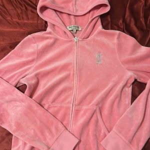Rosa hoodie från Juicy Couture - Säljer en supermjuk och gosig rosa hoodie från Juicy Couture i storlek M. Den har en dragkedja framtill och en snygg JC-logga på bröstet. Perfekt för en chill dag eller när du vill känna dig extra bekväm. 💖