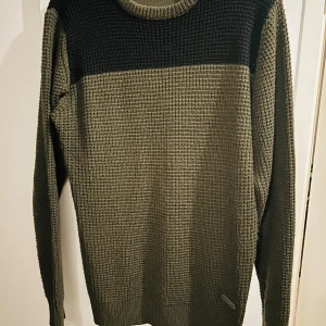 Olivgrön stickad tröja från Brave Soul - Säljer en snygg olivgrön stickad tröja från Brave Soul med svart panel upptill. Tröjan har en klassisk rund hals och långa ärmar, perfekt för kyliga dagar. Den är i mycket bra skick och passar både till vardags och lite finare tillfällen. 🖤