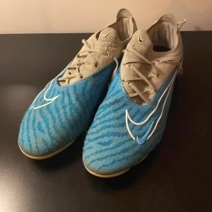 Blå fotbollsskor från Nike - Säljer ett par blå fotbollsskor från Nike i bra skick. Skorna har ett coolt mönster och är utrustade med snörning för perfekt passform. De har en vit sula med dobbar, perfekt för konstgräsplaner. Skorna är både snygga och funktionella, redo för nästa match! Skorna är AG. Skorna är specialdesignade från Nikes hemsida, elite modellen vilket motsvarar den bästa.