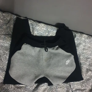 Grå och svarta mjukisbyxor från Nike - Säljer ett par bekväma mjukisbyxor från Nike i grått och svart. De har en elastisk midja med snörning och den klassiska Nike-loggan på sidan. Perfekta för en avslappnad dag hemma eller lätt träning.