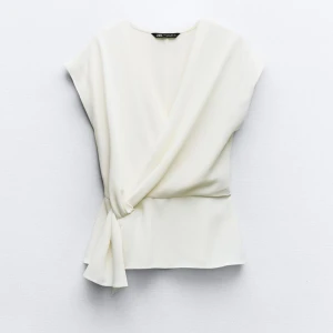 Vit omlottopp från Zara - Säljer en superfin vit omlottopp från Zara. Den har en elegant knytning i sidan och är ärmlös, vilket gör den perfekt för både vardag och fest. Materialet känns mjukt och lyxigt, och toppen har en stilren design som passar till allt. Aldrig använd❤️