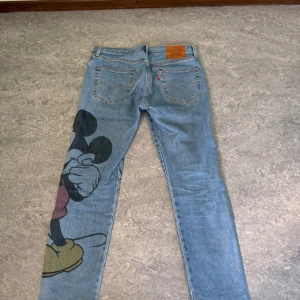 Jeans med Musse Pigg-motiv - Säljer ett par coola jeans från Levis (502-modell) med ett stort Musse Pigg-tryck på ena benet. De är ljusblå och har en klassisk femficksdesign. Perfekta för dig som vill sticka ut lite extra! 🐭👖