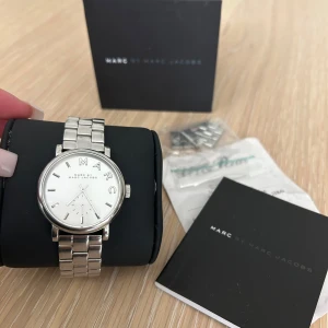 Silverfärgad klocka från Marc Jacobs - Säljer en elegant silverfärgad klocka från Marc Jacobs i mycket bra skick. Klockan har en stilren design med en vit urtavla och silverarmband. Perfekt för både vardag och fest! Extra länkar medföljer för justering av storlek. Batterier behöver bytas, enkelt och billigt att fixa. Små små repor runt silver armbandet som knappt är märkbara.