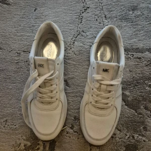 Vita sneakers från Michael Kors - Säljer ett par stilrena vita sneakers från Michael Kors i mycket bra skick. Skorna har en snygg silverdetalj vid hälen och klassisk snörning. Perfekta för både vardag och festliga tillfällen. Storlek och passform är normal.