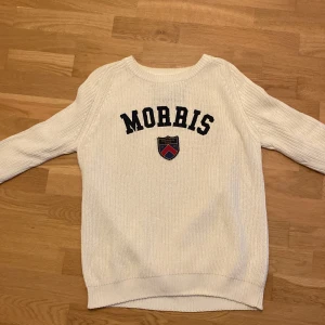 Vit stickad tröja från Morris - Säljer en stilren vit stickad tröja från Morris med text och emblem på framsidan. Tröjan har långa ärmar och en rund halsringning. Perfekt för en klassisk och avslappnad look.