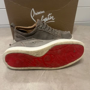 Grå sneakers från Christian Louboutin - Snygga grå sneakers från Christian Louboutin med den ikoniska röda sulan. Skorna har snörning och är tillverkade i ett stilrent material som ger en elegant look.