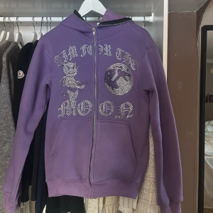 Aim for the moon tröja  - Cool lila hoodie från Aim for the Moon med strassdetaljer som bildar text och motiv på framsidan. Tröjan har en dragkedja och är perfekt för en avslappnad stil. Den har långa ärmar och en mysig huva. Dragkedjan går att knäppa hela vägen upp över luvan! Inga rhinestones har lossnat 