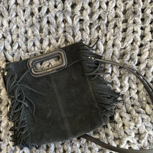 Svart axelväska med fransar från Maje - Snygg svart axelväska i mocka från Maje med coola fransdetaljer. Väskan har ett praktiskt handtag och en avtagbar axelrem, perfekt för en trendig look.