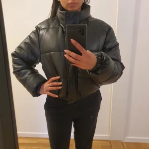 Svart pufferjacka från Zara - Snygg svart skinn pufferjacka från Zara i stl xs, men går att använda som stl.S också.. Jackan har en quiltad design med dragkedja och knappar framtill. Perfekt för kyligare dagar med sin vadderade stil och långa ärmar. Ny pris 1200kr