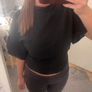 Svart ribbad topp med knyt där bak - Snygg svart ribbad topp med korta ärmar och öppen rygg. Toppen har en unik design med knytning i ryggen som ger en trendig look. Perfekt för att skapa en stilren outfit.