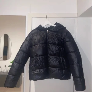 Svart pufferjacka med huva - Säljer en svart pufferjacka i mycket bra skick. Den har en dragkedja framtill och en mysig huva. Perfekt för kalla vinterdagar och höstpromenader. Jackan är långärmad och har en glansig finish som ger en cool look. Passar både till vardags och lite mer uppklädda tillfällen.