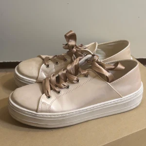 Beige sneakers med satinband - Snygga beige sneakers med breda satinband som snörning. Skorna har en stilren design med en tjock vit sula som ger extra komfort. Perfekta för en avslappnad och trendig look.