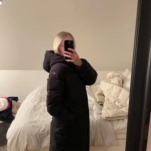 Svart dunjacka med huva - Säljer en svart lång dunjacka från Five seasons, köpt förra vintern. Är en strl 34 och passar mig som är både XS/S. Går ner till knäna (är 161 cm lång). Snöret man hänger jackan i är sönder och en liten blekt fläck finns på jackan längst ner till vänster. Vita prickar är pga smutsig spegel. 💕 