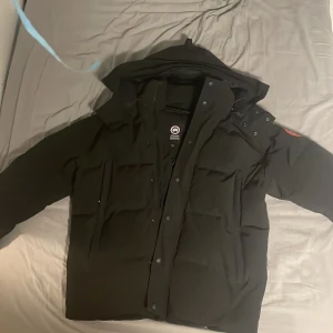 Svart dunjacka från Canada Goose - Säljer en svart dunjacka från Canada Goose med broderad logga på ärmen. Jackan har en huva och stängs med dragkedja och knappar. Perfekt för kalla vinterdagar. Använd runt 8 gånger. Finns även en scanner som leder till canada goose original sida. 