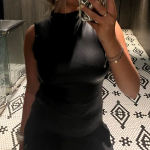 Svart ärmlös topp - Snygg svart ärmlös topp i ett glansigt material. Perfekt för en kväll ute eller en stilren look. Toppen har en hög hals och en tight passform som framhäver figuren.