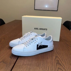 Vita sneakers från Axel Arigato - Snygga vita sneakers från Axel Arigato med svart fågelbroderi på sidan. Box och original tillbehör medföljer, nypris ungefär 2500kr. Hör av dig vid frågor! 