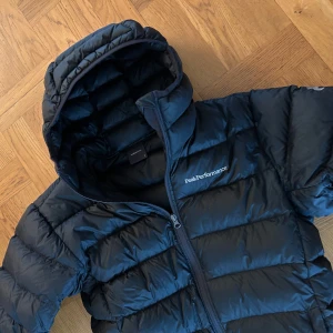 Svart dunjacka från Peak Performance - Snygg och varm svart dunjacka från Peak Performance med huva och dragkedja. Perfekt för kyliga dagar med sin quiltade design som håller värmen inne.