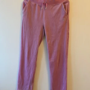 Rosa mjukisbyxor från Juicy Couture - Supermysiga rosa mjukisbyxor från Juicy Couture i velourmaterial. De har en bekväm passform med resår och snörning i midjan. På baksidan finns den ikoniska 'Juicy'-texten i glittriga stenar. Perfekta för en chill dag hemma eller en avslappnad utflykt. 🩷