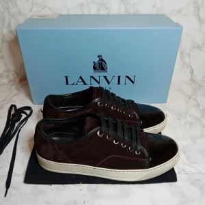 Lanvin skor storlek 40 - Snygga bruna sneakers från Lanvin med svarta detaljer och snörning de är i storlek uk5. Skorna har en stilren design med en kombination av mocka och glansigt material på tån. Perfekta för en trendig look. Box ingår inte