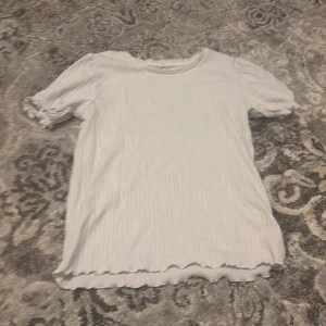 Vit ribbad topp - Säljer en söt vit ribbad topp med korta puffärmar och volangdetaljer vid ärmslut och nederkant. Perfekt för en avslappnad stil.