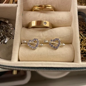 Söta hjärtformade örhängen med lila stenar - Säljer ett par supersöta örhängen i form av hjärtan. De är med glittriga ljusblåa stenar och har en pilformad detalj. De är köpta i Spanien så vet inte riktigt vilket märke det är!💕