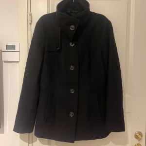 Svart kappa med knappar - Säljer en stilren svart kappa i mycket bra skick. Den har långa ärmar och stängs med stora knappar framtill. Perfekt för höst och vinter, och ger en elegant look. Passar bra till både vardag och fest!
