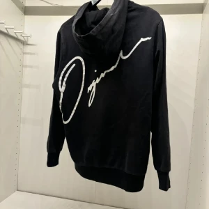 Svart hoodie med vit text - Säljer en svart hoodie med en stor vit signaturtext på ryggen. Den har en klassisk passform med långa ärmar och en bekväm huva. Perfekt för en avslappnad stil eller kyliga dagar. Passar både till vardags och chillkvällar. Strl S/M S men sitter som M
