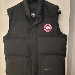 Svart dunväst från Canada Goose - Säljer en svart dunväst från Canada Goose i mycket bra skick. Västen har en klassisk design med knappar och dragkedja framtill samt en broderad logotyp på bröstet. Perfekt för kyliga höst- och vinterdagar. Passar både till vardags och mer uppklädda tillfällen.