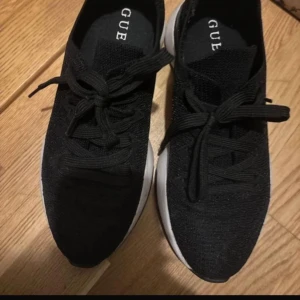 Svarta sneakers från Guess - Säljer ett par snygga svarta sneakers från Guess i storlek 37. De är i bra skick och har en stilren design med snörning. Perfekta för både vardag och träning. Sulan är vit vilket ger en cool kontrast till det svarta tyget. Passar perfekt till alla outfits!