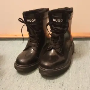 Snygga svarta boots från Hugo i blankt vattenavvisande material. De har snörning framtill och en mysig foderkant som håller dig varm. De är fodrade invändigt i hela skon. Perfekta för höst och vinter, både stiliga och praktiska. Passar till både jeans och kostymbyxor! 