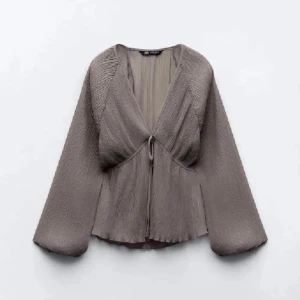 Grå blus från Zara - Elegant grå blus från Zara med v-ringad design och knytning framtill. Blusen har långa, vida ärmar och ett lätt plisserat material som ger en fin struktur. Fint skick förutom att en pyttelitentråd gått upp där bak tror man kan klippa eller sy. Kan skicka bild vid intresse💕
